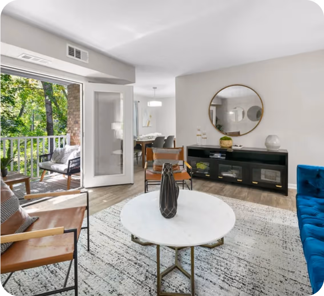 Modern living room with open door to a balcony, featuring a blue sofa, leather chairs, round white coffee table, and a round mirror above a black console.